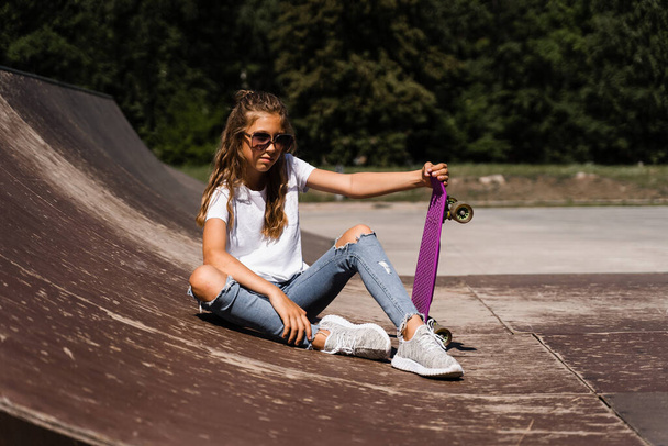 Child model in glasses sitting and posing with penny board on skate playground. Active child girl with enjoy extreme lifestyle on sport ramp - 写真・画像
