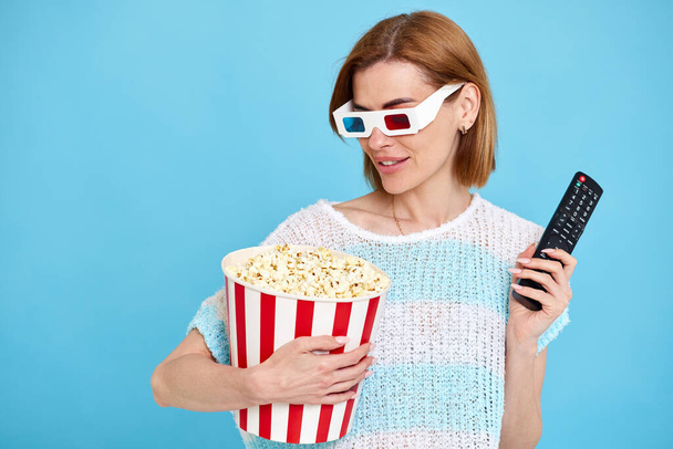 beautiful woman in 3d glasses with remote control watching movie film and holding bucket of popcorn on blue background. copy space - Foto, imagen