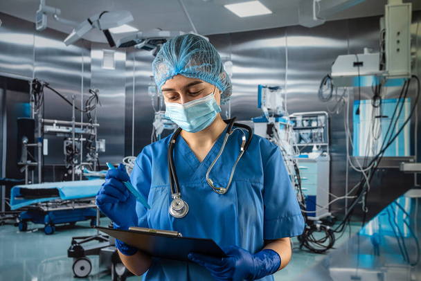 Female doctor in blue uniform and protective mask, gloves writes notes on the clipboard in operating room at hospital - Foto, Imagem