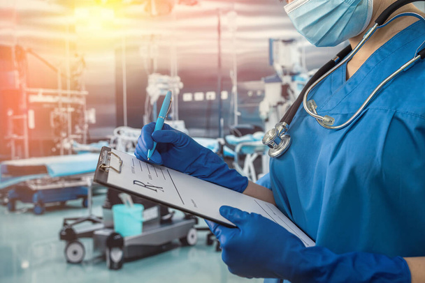 Ärztin in blauer Uniform und Schutzmaske, Handschuhe schreiben Notizen auf Klemmbrett im Operationssaal des Krankenhauses - Foto, Bild