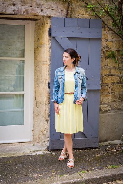 Lifestyle portrait of young stylish woman with long brunette hair walking on the street in old town, wearing yellow dress and denim jacket - Photo, Image
