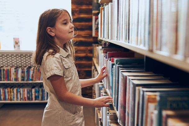 Schoolgirl choosing book in school library. Smart girl selecting books. Books on shelves in bookstore. Learning from books. School education. Benefits of everyday reading. Child curiosity. Back to school - Φωτογραφία, εικόνα