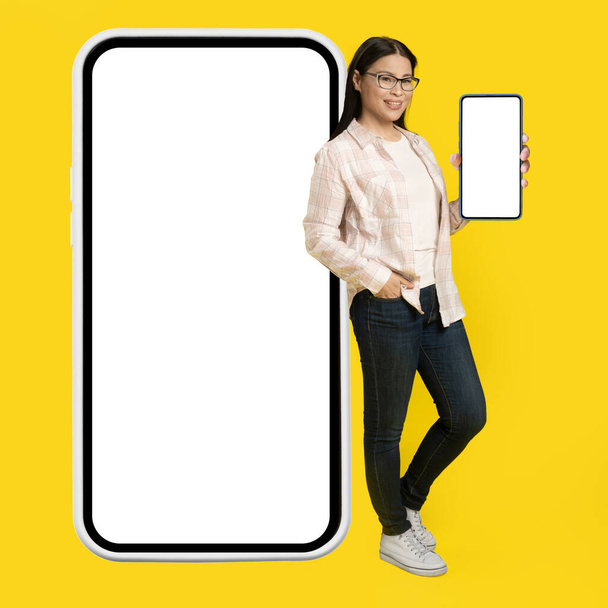 Showing white screen on phone charming middle aged asian woman leaned back on giant, huge smartphone with white screen wearing casual isolated on yellow background. Free space mock up. - Photo, Image