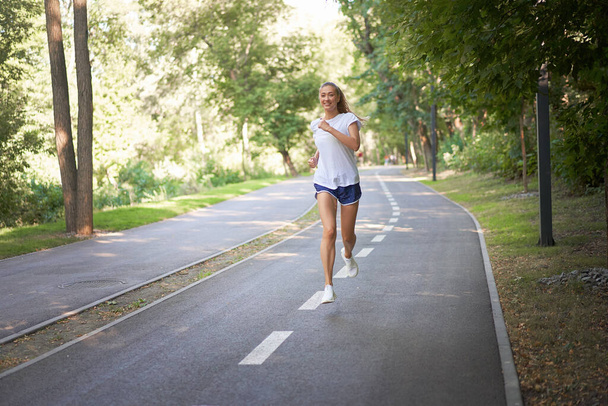 Woman running asphalt road summer park Active sporty caucasian female morning workout Healthy lifestyle concept. Athletic person sportswear exercises Dressed white t-shirt full length - Φωτογραφία, εικόνα