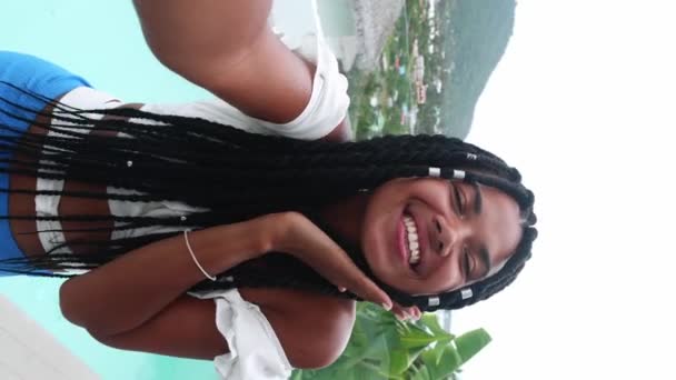 Cute woman shows peace sign, blows air kiss and cutely smiles looking at web cam in smartphone, against tropic landscape - Video