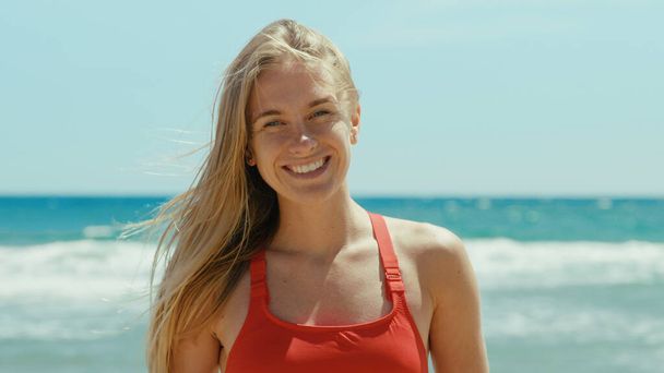 Portrait of beautiful young woman in red swimsuit stand on empty sunny beach, look into camera with confidence. Happy authentic smile. Summertime concept. Warm breeze in hair - Photo, Image