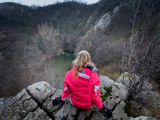 Woman sitting down on top of a cliff and looking down into the valley at a flowing river - Foto, Imagen