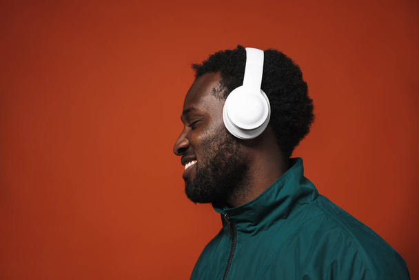 Black bristle man smiling while listening music with headphones isolated over brown wall - Foto, afbeelding