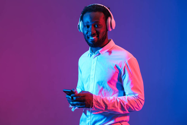 Mor duvarda izole edilmiş kulaklık ve cep telefonuyla müzik dinleyen genç bir zenci. - Fotoğraf, Görsel
