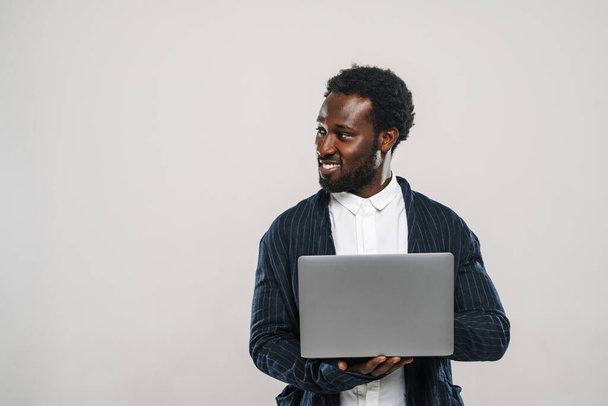 Young black man wearing jacket smiling while typing on laptop isolated over white wall - Φωτογραφία, εικόνα