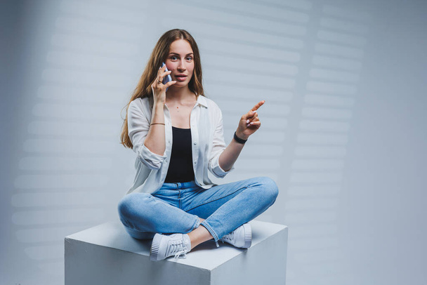Young woman with long brunette hair, wearing a white shirt and jeans, talking on the phone. A woman in jeans and a plain white shirt with a mobile phone. White background, copy space. - Foto, Imagem