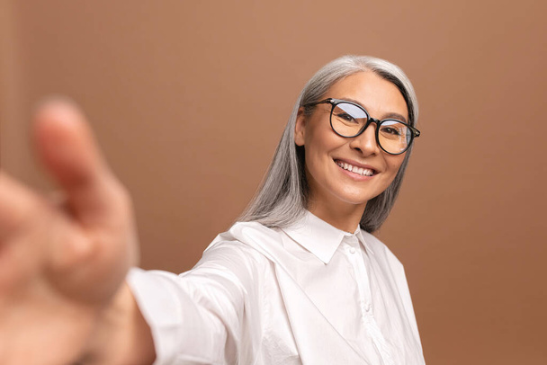 Lady wearing glasses smiling and looks at the camera isolated on beige. Cheerful senior asian woman taking selfie. POV - Photo, image