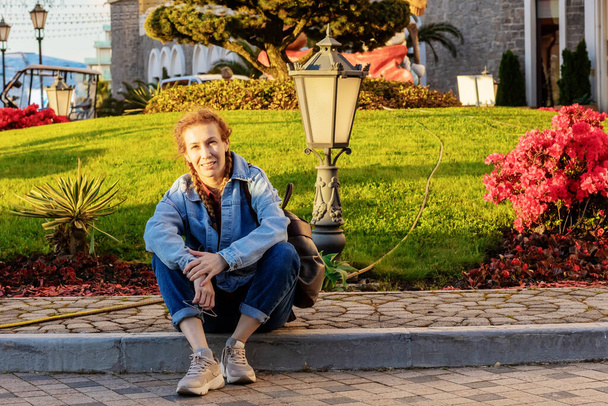 Eine Frau im Jeans-Outfit sitzt auf einem Steinweg in der Nähe eines grünen Rasens mit südlichen Pflanzen, Blumen und Laternen im Sonnenuntergang. Touristischer Spaziergang. - Foto, Bild