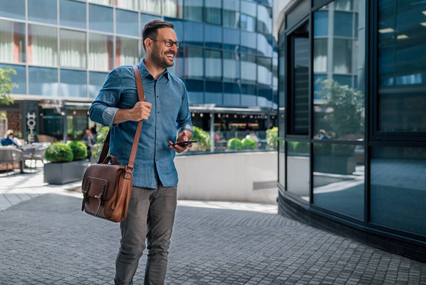 Happy entrepreneur walking with smart phone. Young adult professional wearing casuals walking in business quarters. He is going on footpath against business buildings during sunny day. - Fotografie, Obrázek