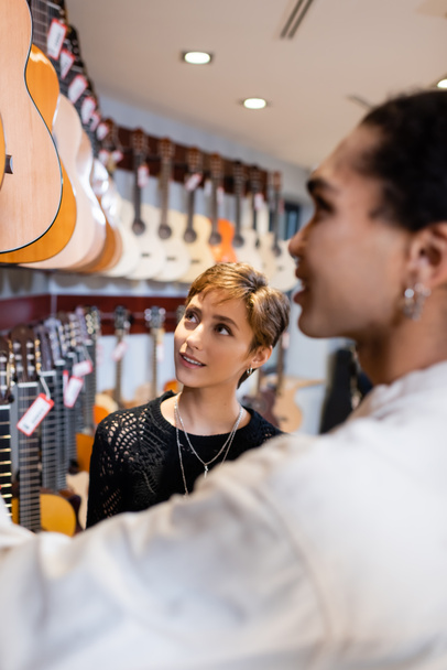 Улыбающийся покупатель смотрит на акустические гитары рядом с размытым африканским продавцом в музыкальном магазине  - Фото, изображение