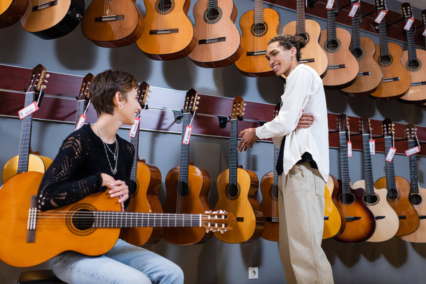 Enstrümantal müzik mağazasında Afrika kökenli Amerikalı bir satıcının yanında akustik gitar tutan gülümseyen bir kadın.  - Fotoğraf, Görsel