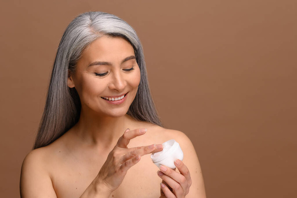 Charming good-looking Asian middle-aged woman with naked shoulders, looks down at jar of anti-aging moisturiser in her hand isolated beige background. Spa and relax, cosmetology procedure - Foto, immagini