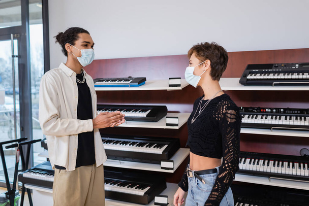 African american seller in medical mask looking at customer near synthesizers in music store  - Fotografie, Obrázek