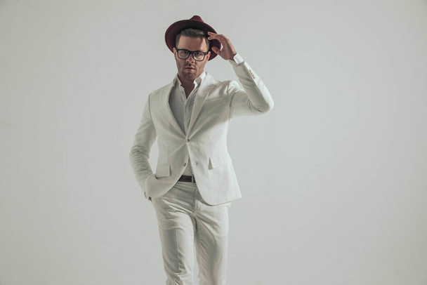 sexy hombre de negocios en traje blanco con la mano en el bolsillo que arregla el sombrero y se mueve delante del fondo gris en el estudio - Foto, Imagen