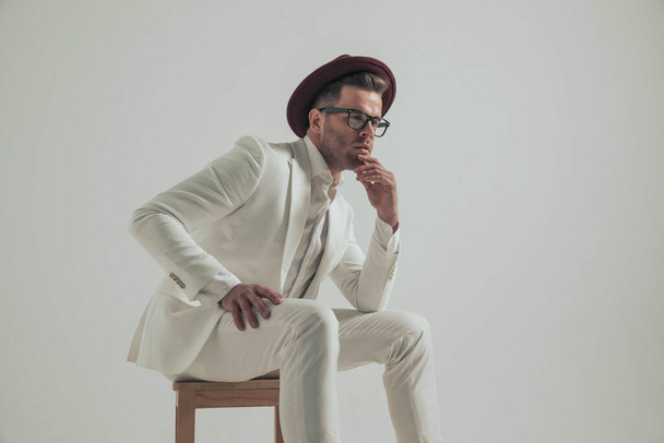 zamyślony młody człowiek w eleganckim stroju casual trzymając rękę do podbródka i myśląc siedząc na drewnianym krześle na szarym tle w studio - Zdjęcie, obraz