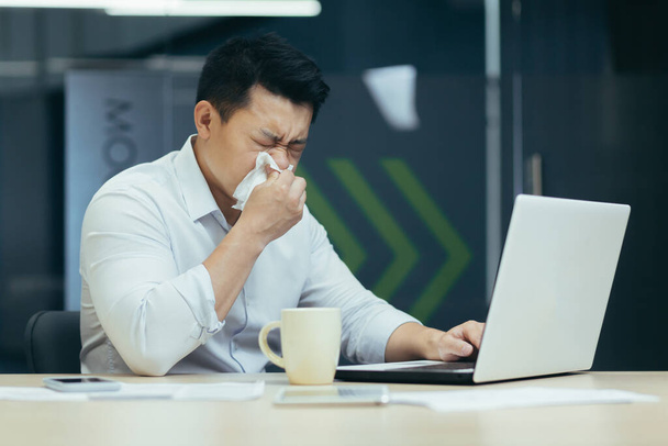 Sick man in office coughs and has runny nose, Asian businessman with cold works in office on laptop - Foto, Bild