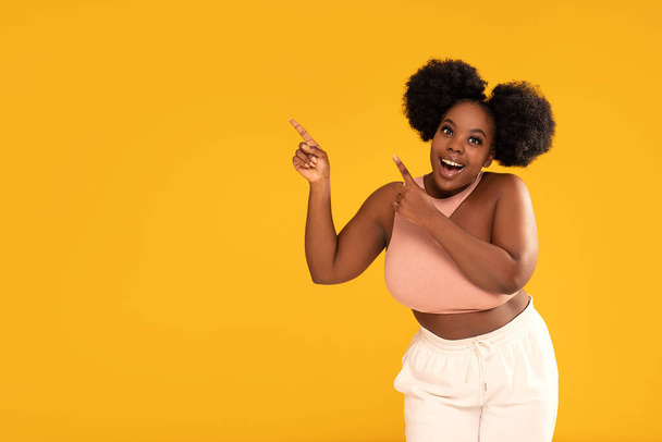 Happy smiling woman pointing at copy space on yellow studio background. Young beautiful african girl showing something on empty space. Conceptual photo. - Foto, afbeelding