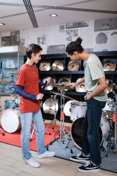 Vista lateral del vendedor sosteniendo palos de tambor cerca de cliente afroamericano y tambores en tienda de música  - Foto, imagen