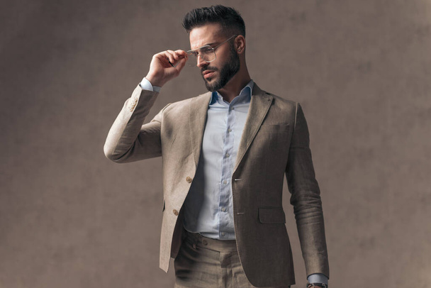 portrait of elegant unshaved businessman with undone shirt looking to side and adjusting glasses in front of beige background in studio - Fotó, kép