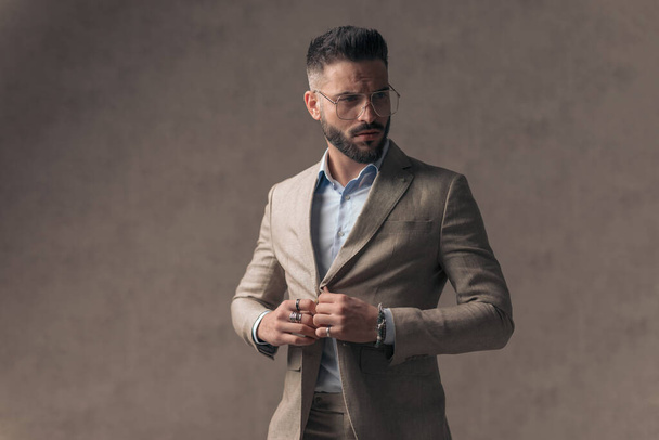 portrait of sexy bearded businessman with open collar shirt looking to side and adjusting suit, posing in front of beige background in studio - Φωτογραφία, εικόνα