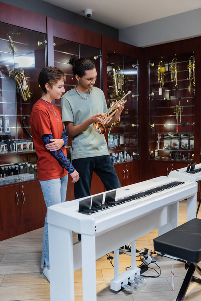 Interracial seller and customer looking at trumpet near piano in musical store  - Φωτογραφία, εικόνα