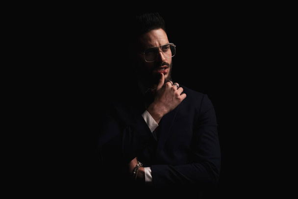 irresistible elegant man in suit looking to side and sensually touching lips while posing with studio lights in front of black background in studio - Фото, зображення
