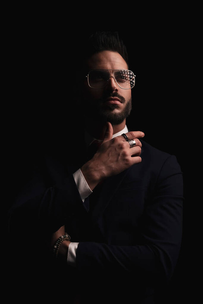 confident businessman in elegant suit crossing arms and looking away on black background in studio - Fotó, kép