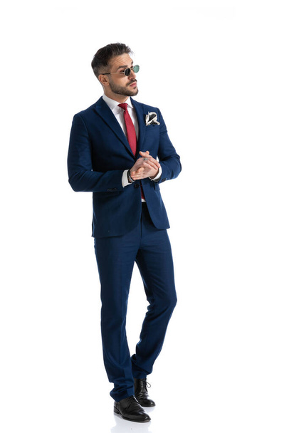 handsome unshaved man in suit holding hands together and looking to side, walking and posing in front of white background in studio - Photo, Image