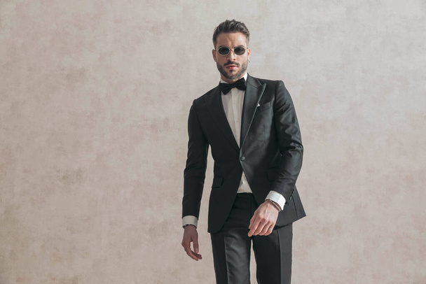 sexy bearded groom with sunglasses being cool while walking, wearing black tuxedo with black tie in front of beige background in studio - Photo, Image