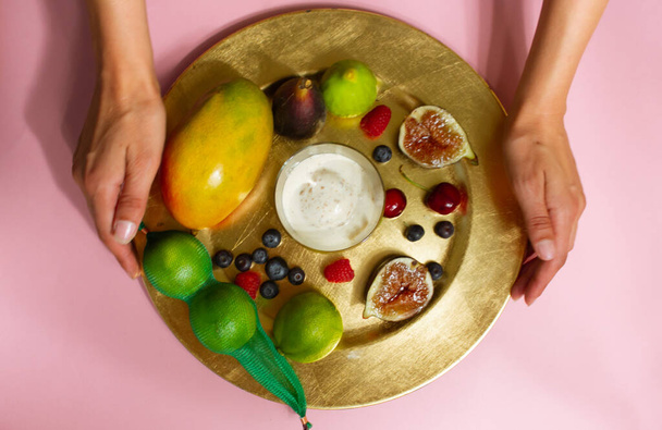 A női kezek egy gyönyörűen tálalt tányért, friss trópusi gyümölcsöt szolgálnak fel rózsaszín asztallapon. Arctalan lány az asztalon. Füge, mangó, bogyók, cseresznye, lime, jégkrém tálcán - Fotó, kép
