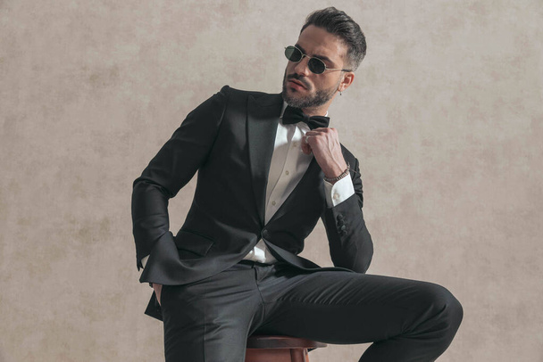 portrait of cool young groom in black tuxedo looking to side and fixing bowtie, holding hand in pocket and sitting on wooden chair on beige background in studio - Foto, Imagen