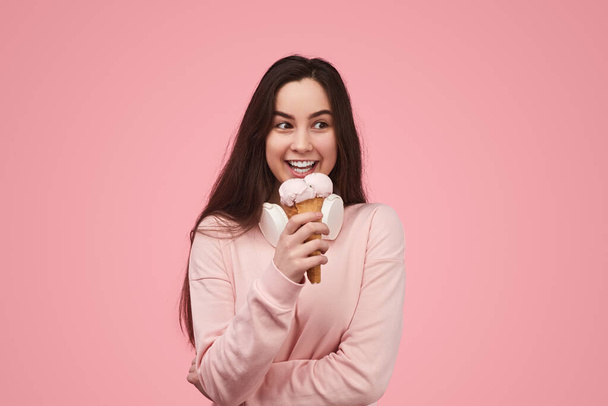 Cheerful young lady with long dark hair in sweatshirt and headphones on light, smiling and looking away with curiosity while eating delicious ice cream cone against pink background - Photo, Image