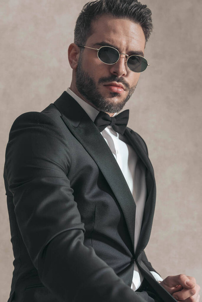 cutout picture of cool elegant groom in black tuxedo with sunglasses posing in front of beige background in studio - Foto, imagen