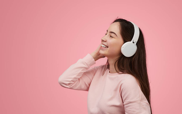 Cheerful teen meloman in sweatshirt touching wireless headphones and smiling with closed eyes while enjoying good music against pink background - Photo, image