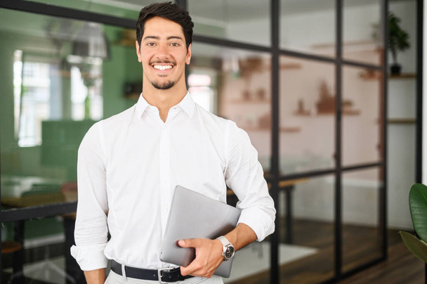 Working with joy. Smiling cheerful hispanic business man wearing formal white shirt standing with laptop computer in office, looking at camera, proud and ambitious latin start-up owner - Foto, Imagem