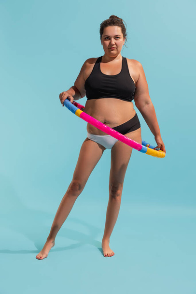 Motivation to be beautiful and healthy. Young plus-size woman with slender legs doing exercises with hula hoop isolated on colored background. Weight loss, fitness, healthy eating, active life - Zdjęcie, obraz