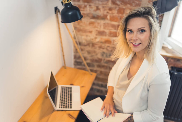 Professional caucasian pregnant businesswoman in a white blazer looking at camera while sitting at her office desk with a laptop and a calendar. High quality photo - Foto, Imagem
