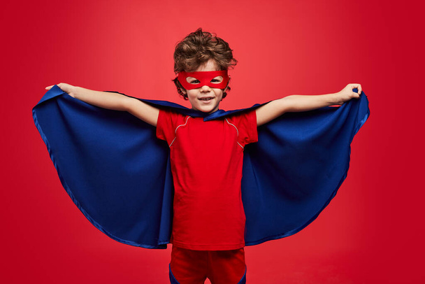 Glad little boy in superhero costume looking at camera with smile and spreading cloak while playing against red background - Foto, Imagem