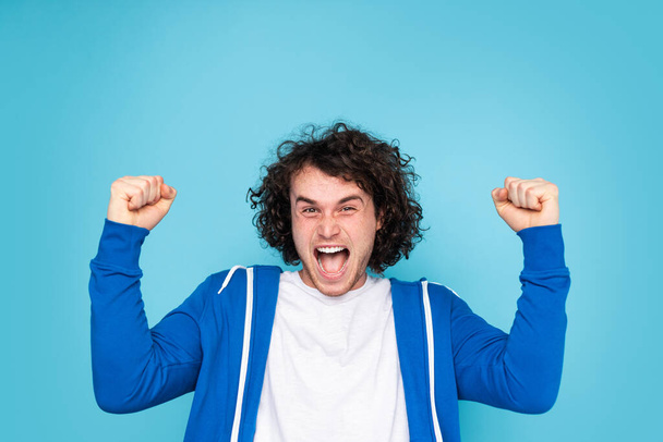 Excited young male with curly dark hair in casual clothes clenching fists and screaming happily, while looking at camera and celebrating success against blue background - Photo, image