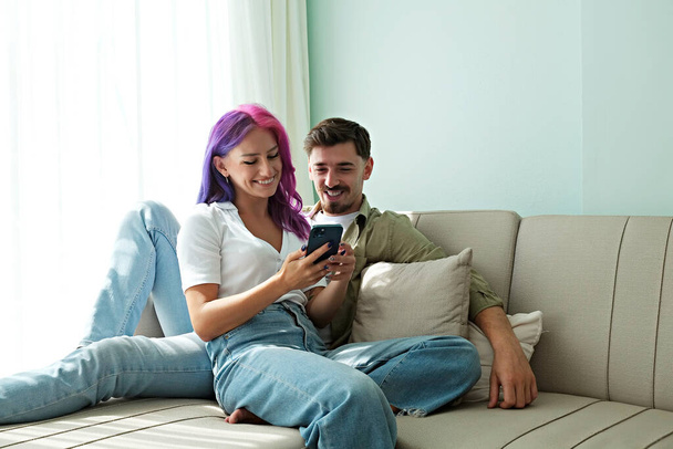 Hipster couple sitting on the couch laughing at memes on the phone at home. Young woman with bright colorful hair and bearded man in the living room having fun. Close up, copy space, background - Foto, Bild