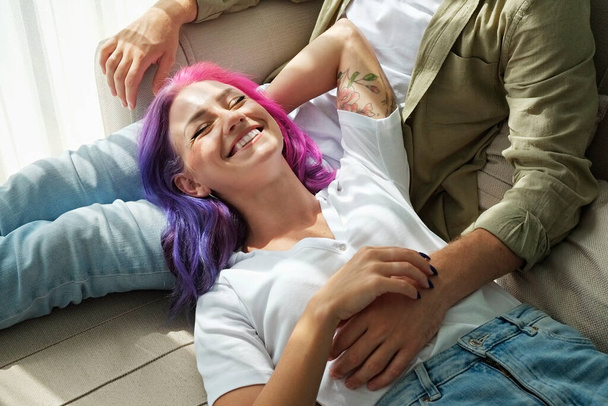 Hipster couple hugging on the couch at home. Tattooed young woman with bright colorful hair and her man relaxing in the living room. Close up, copy space, background. - Foto, Imagem