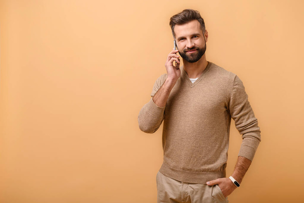 Young bearded man talking phone, calling to somebody, looking at the camera with happy facial expression. Indoor studio shot isolated on beige background. - Foto, imagen