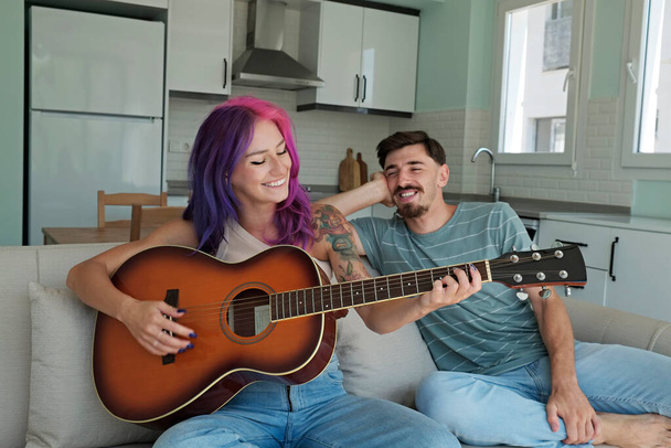 Young beautiful woman with colorful pink and purple hair playing a song on the guitar for her boyfriend. Tattooed female and bearded male having fun singing songs. Close up, copy space, background. - Photo, image