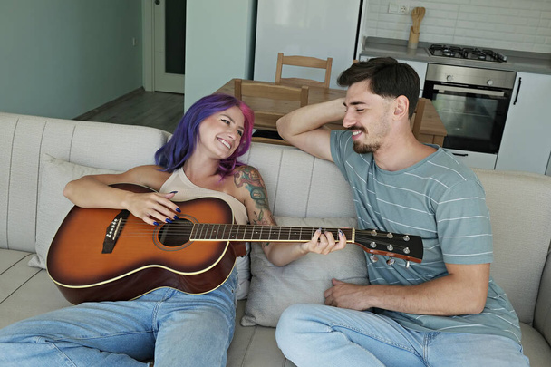 Young beautiful woman with colorful pink and purple hair playing a song on the guitar for her boyfriend. Tattooed female and bearded male having fun singing songs. Close up, copy space, background. - Φωτογραφία, εικόνα
