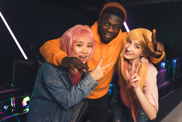 Multiracial young cheerful friends standing close together posing at camera in a dark gaming room with neon lights showing peace and thumbs up signs. High quality photo - Foto, imagen